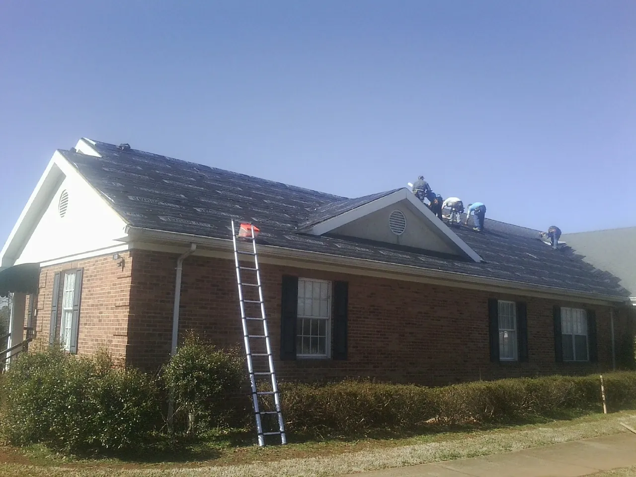 Crew tearing shingles off a brick ranch roof with a ladder set — Metal Roof Repair work by our crew in Shacklefords
