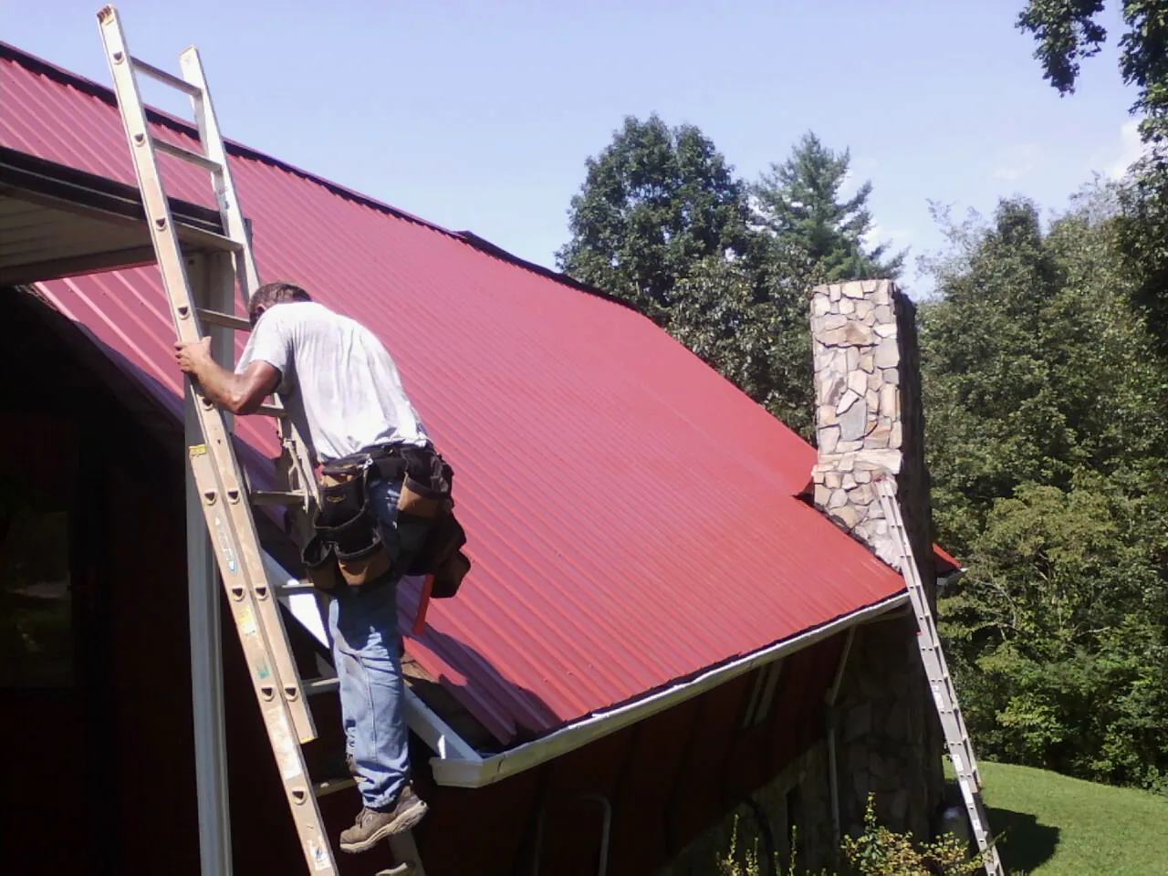 Roofer on a ladder beside a red metal roof and stone chimney — Soffit & Fascia Repair by Roofing Brothers in Shacklefords